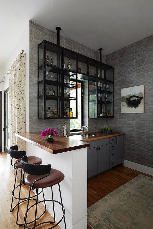 Chic metal and glass French bar shelves are mounted to a gray wall around a window and over gray flat front cabinets. The cabinets don oil rubbed bronze hardware and a wooden countertop holding a small round metal sink beneath a polished nickel faucet Brown leather and metal counter stools sit at a white island finished with a wood top.