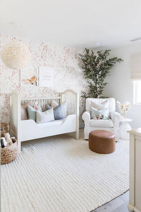 White and pink girl's nursery features a pink accent wall with a white French toddler bed atop a cream knitted rug, illuminated by a white feather ball chandelier and a white slipcovered nursery glider with a round leather ottoman.