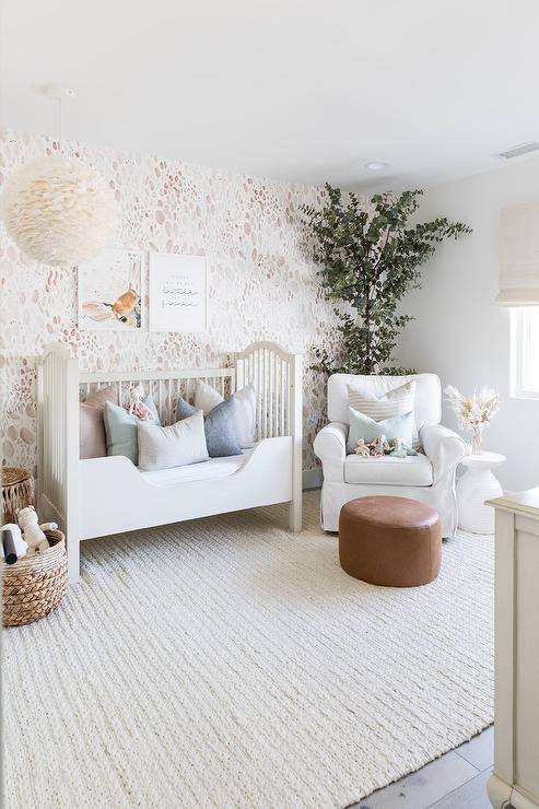 White and pink girl's nursery features a pink vocalizing wall with a white French toddler bed atop a surf knitted rug, illuminated by a white feather wittiness chandelier and a white slipcovered nursery glider with a round leather ottoman.