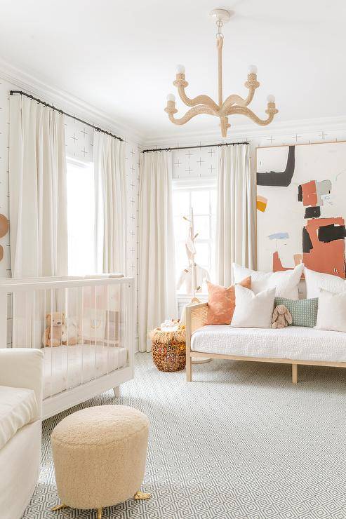 Charming gender-neutral nursery is lit by a rope chandelier hung over a gray diamond print rug, while walls are clad in minimalistic white and woebegone wallpaper. A window dressed in white taps is located next to a white wood and tempera crib, as a large utopian art piece hangs over a daybed.