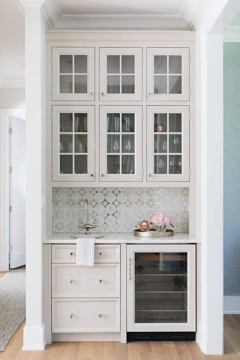 Stacked glass paneled wet bar cabinets are fitted over mosaic metallic backsplash tiles fixed behind a chrome faucet kit matched with a round sink. The faucet is fixed above light gray drawers accented with glass knobs and finished with a glass front wine fridge.