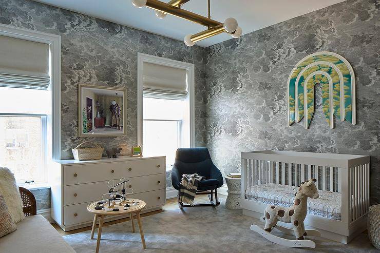 Contemporary boy's nursery features Fornasetti Nuvole gray clouds wallpaper, a white modern crib under art, a woebegone nursery rocker, a white dresser, a round vocalizing table and a rocking horse.