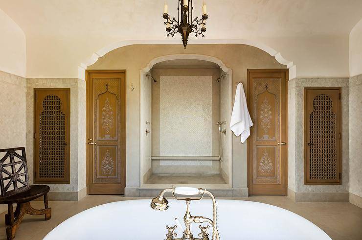 Moroccan style master bathroom features an open shower clad in ivory and cream tiles framing marble mosaic accent tiles and flanked by Moroccan style doors.