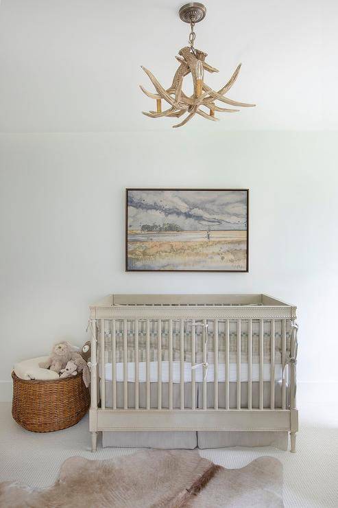 A tan cowhide rug sits on off-white carpet in front of a gray wooden crib accented with a gray crib skirt and placed underneath a framed landscape art piece. The room is illuminated by a faux antlers chandelier.