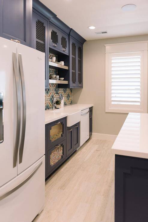 Basement kitchenette features blue cabinets paired with glossy white quartz countertops and a tan and green mosaic tiled backsplash. Basement kitchen boasts metal lattice upper cabinets flanking stacked stainless steel shelves over a farm sink with clear see through candy drawers to the left and a white dishwasher to the right.