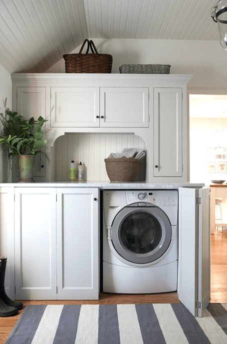 Amazing mudroom features white beadboard ceiling over white shaker cabinets paired with white marble countertops and beadboard backsplash. Cottage mudroom with concealed washer and dryer hidden behind folding doors over white and gray striped rug.