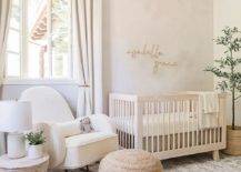 A soft pale pink boho chic nursery features gold letters on pale pink wallpaper over a light beige wooden nursery crib and a white boucle nursery rocker with tan ottoman and round accent table on white and gray layered rugs.