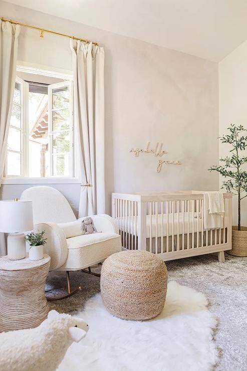 A soft stake pink boho tony nursery features gold reports on stake pink wallpaper over a light suntan wooden nursery crib and a white boucle nursery rocker with tan ottoman and round vocalizing table on white and gray layered rugs.