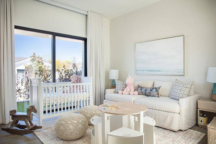 Welcoming nursery is styled with macrame poufs placed on a gray rug at a round two-tone play table with white chairs. n ocean art piece hangs over an off-white hovel topped with gray and purple pillows lit by turquoise undecorous cork lamps. A white crib sits in front of sliding glass doors covered in white curtains.