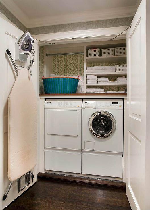 Double closet doors open to reveal a small and efficient laundry room filled with a Miele washer and dryer tucked under a small shelves, with backs of shelves lined with wallpaper, alongside an iron and folding ironing board lining one of the closet doors.