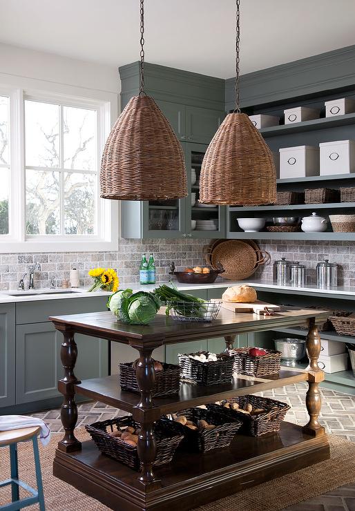 Green cottage kitchen features a brown French freestanding center island illuminated with basket chandeliers. Perimeter green cabinets feature open shelving with gray brick backsplash tiles complimenting gray brick herringbone floor pavers.
