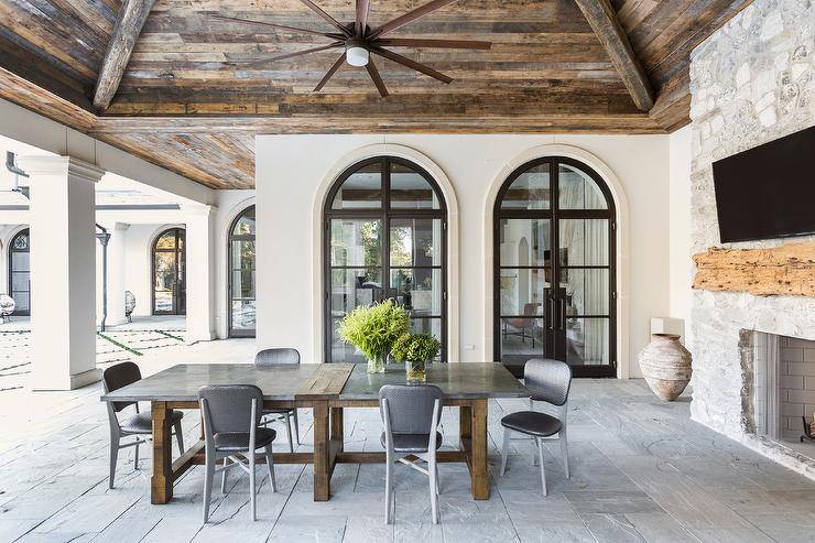 A vaulted rustic wood plank covered patio ceiling holds a ceiling fan over a long metal and wood dining table surrounded by black chevron dining chairs.