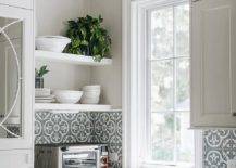 White floating shelves are stacked in a kitchen against a light gray wall and over black and white quaterfoil cement wall tiles fixed partially framing a window. White kitchen cabinets are accented with nickel hardware.
