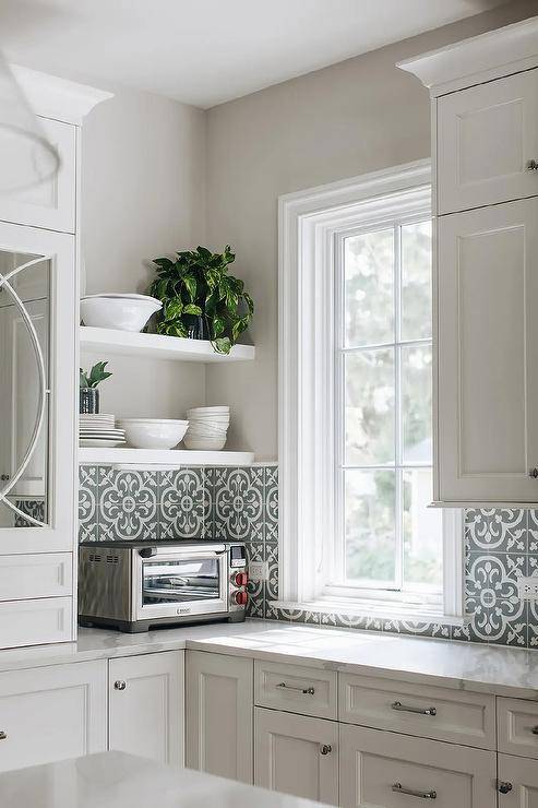 White floating shelves are stacked in a kitchen against a light gray wall and over black and white quaterfoil cement wall tiles fixed partially framing a window. White kitchen cabinets are accented with nickel hardware.