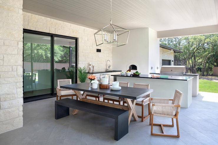 Light gray chairs at an outdoor x base concrete top dining table illuminated by a polished nickel lantern. This outdoor patio is completed with charcoal gray floor tiles, white stone walls, and a white plank ceiling.