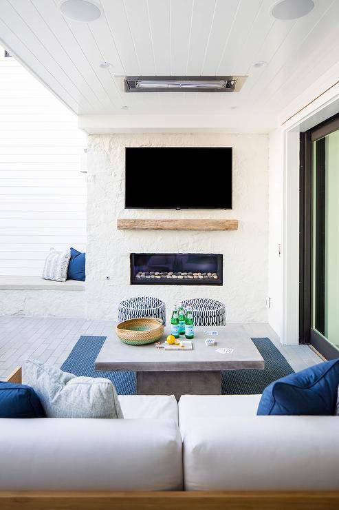Add a ceiling heat lamp to a covered patio for added comfort on a large patio furnished with teak sofas and a concrete coffee table. A wall mount tv is displayed over a white stone fireplace bringing entertainment to an outdoor escape.
