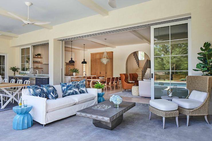 A white slipcovered sofa accented with blue palm print pillows sits on the concrete floor of a covered patio between blue twist stool end tables.