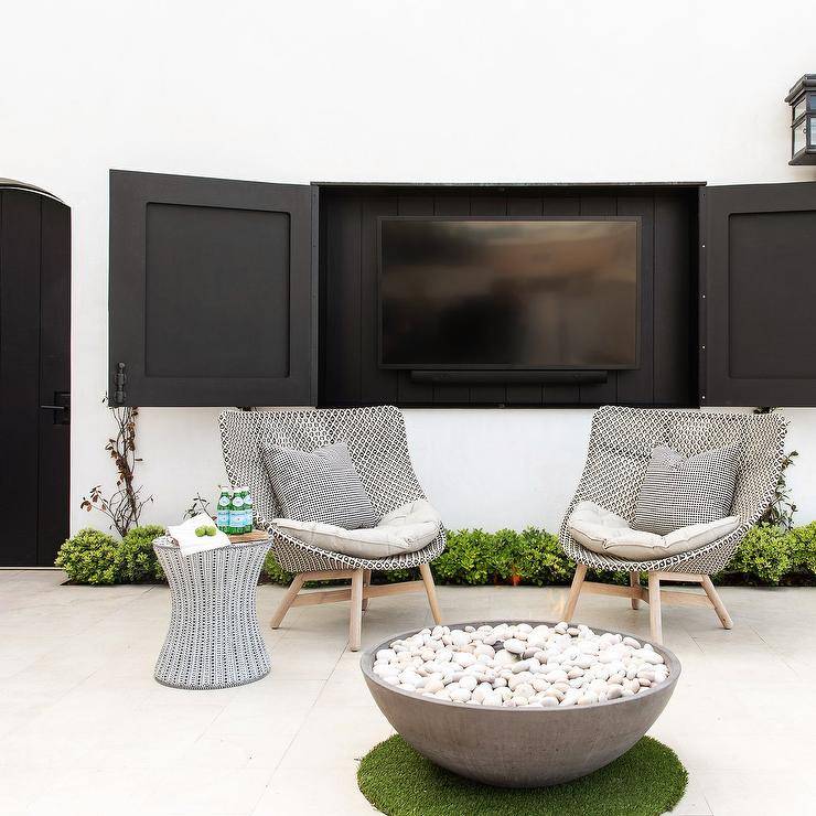 Hidden tv concealed in an outdoor patio with black cabinet doors. The patio is furnished with simplicity yet stylish appeal. A round concrete fire is surrounded by black and white outdoor chairs and a black and white accent table.
