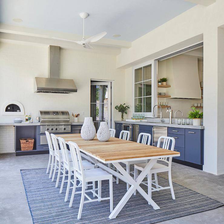 Covered patio dining space features a white trestle dining table fitted with a teak plank top and surrounded by 1006 Navy Chairs placed on a concrete floor covered in a blue rug. Blue outdoor kitchen cabinets are accented with polished nickel hardware and finished with a sink with a polished nickel gooseneck faucet and a stainless steel BBQ fitted beneath a stainless steel range hood.