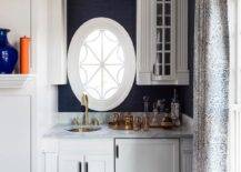 An oval window is framed by blue grasscloth wallpaper and positioned beside glass front cabinets and over white wet bar cabinets. The cabinets are topped with a gray polished marble countertop holding a round brass sink under an antique brass gooseneck fault. The wet bar is finished with a white wood paneled