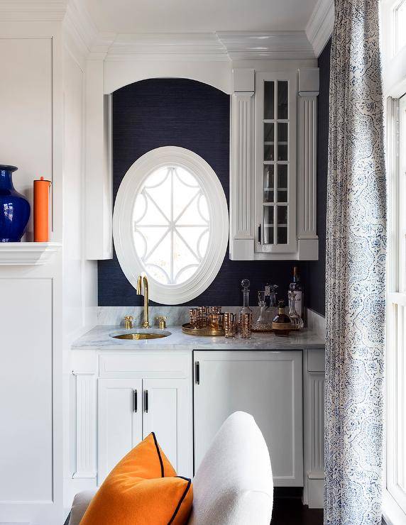 An oval window is framed by blue grasscloth wallpaper and positioned beside glass front cabinets and over white wet bar cabinets. The cabinets are topped with a gray polished marble countertop holding a round brass sink under an antique brass gooseneck fault. The wet bar is finished with a white wood paneled