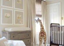 Lovely gender-neutral nursery boasts a black spindle crib placed on a blue arabesque rug beside a large giraffe plush. The plush stands in front of a bamboo roman shade layered under tan colored curtains. Four art prints are framed and hung from a tan wall over a French wooden nursery topped with a changing mat. A blue monogrammed pillow sits atop a tan linen glider.