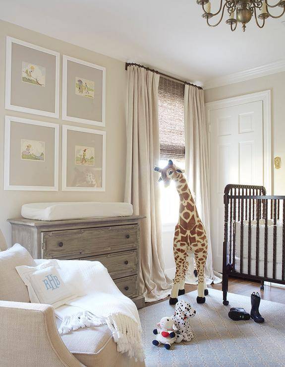 Lovely gender-neutral nursery boasts a black spindle crib placed on a blue arabesque rug beside a large giraffe plush. The plush stands in front of a bamboo roman shade layered under tan colored curtains. Four art prints are framed and hung from a tan wall over a French wooden nursery topped with a changing mat. A blue monogrammed pillow sits atop a tan linen glider.