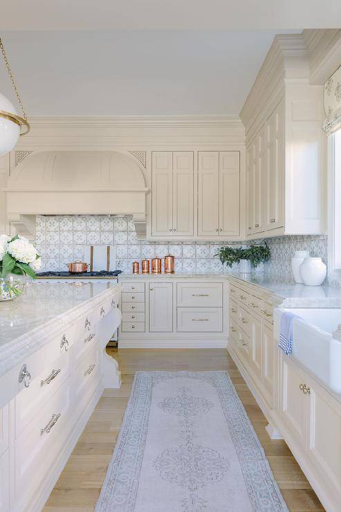 A gray runner accents ivory kitchen cabinets donning satin nickel knobs and topped with a marble countertop fixed against white and gray mosaic wall tiles. Ivory upper cabinets flank an ivory barrel hood mounted above a La Cornue CornuFe range.
