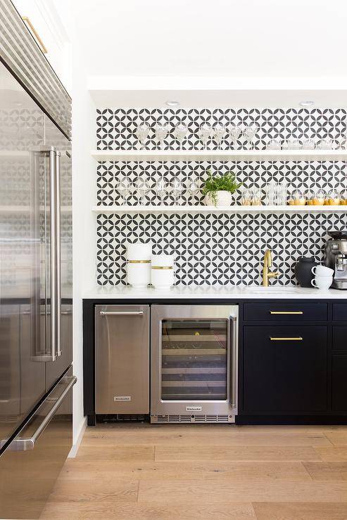 White floating shelves are fixed against black and white backsp[lash tiles in a contemporary wet bar. The shelves are fitted over black flat front cabinets adorned with brass hardware and a white quartz countertop. The countertop is mounted over a Kitchenaid wine fridge and finished with a sink and antique brass faucet.