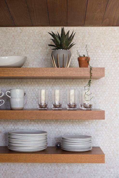 Kitchen features brown floating shelves on white tumbled penny wall tiles.