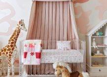 A white and pink canopy hangs from a white and pink wall above a lucite crib matched with pink crib sheets. The crib sits on a gray rug beside a giraffe plush doll.