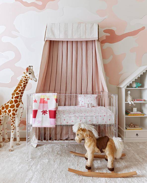 A white and pink canopy hangs from a white and pink wall above a lucite crib matched with pink crib sheets. The crib sits on a gray rug beside a giraffe plush doll.