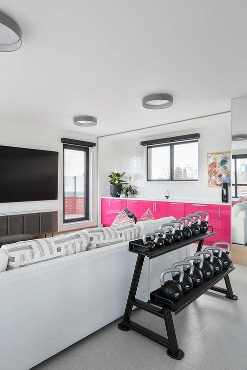Basement gym features a kettle back rack placed behind a white sofa topped with white and gray cushions and positioned facing a wall mount TV. pink cupboards kitchenette
