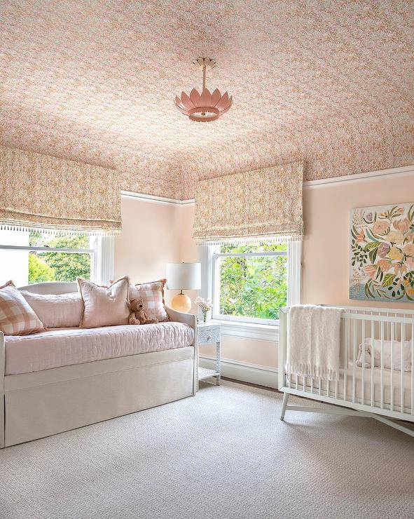 Lovely pink nursery is lit by a pink lotus flower chandelier hung from a pink wallpapered ceiling matching pink roman shades. A white mid-century modern crib sits beneath a floral art piece hung from a pink wall. A light tan skirted daybed sits inf ront of a window and is complemented with pink buffalo check pillows and light pink bedding.