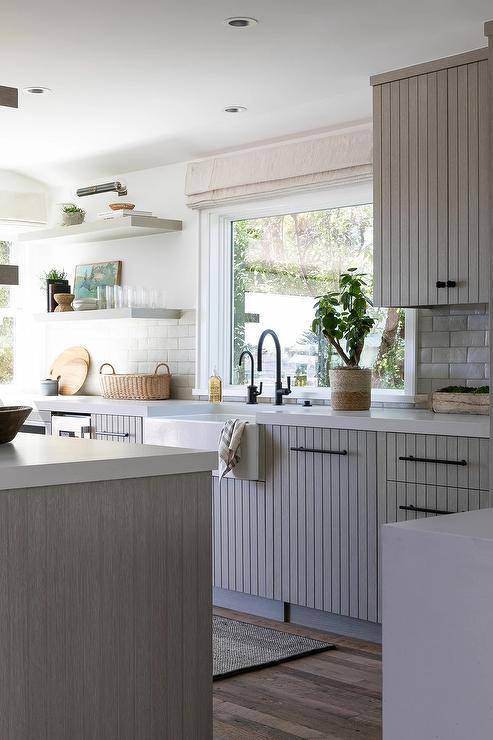 Gray floating shelves and gray plank cabinets adorned with oil rubbed knobs flank a kitchen picture window covered in a gray roman shade hung above a farm sink with an oil rubbed bronze gooseneck faucet. Gray plank lower cabinets are finished with oil rubbed bronze pulls and a white quartz countertop mounted against glazed gray offset backsplash tiles.