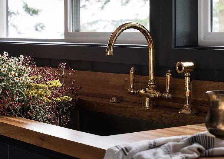 A polished brass vintage faucet is fixed to a wooden countertop beneath a window and over a kitchen sink.