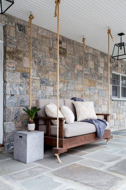 Hung from a beadboard patio ceiling, a wood and rope hanging sofa is positioned beside a concrete end table.