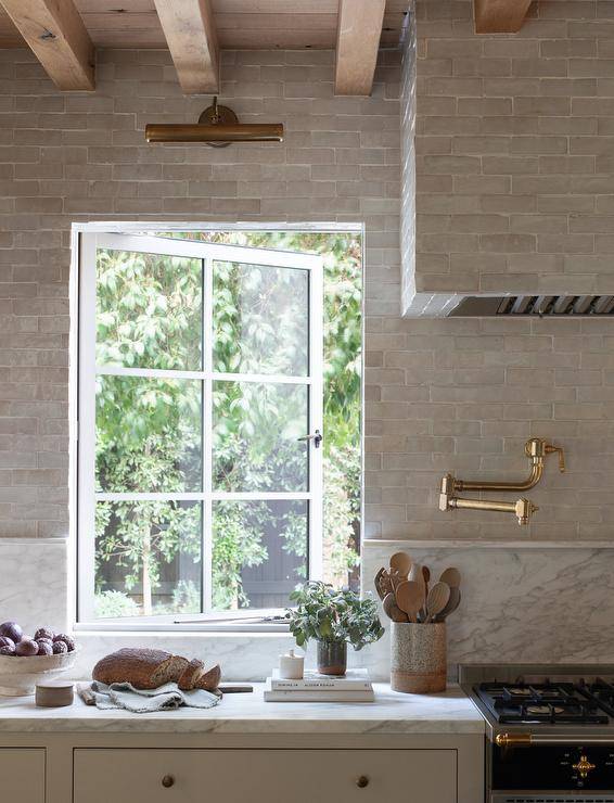 A brass picture light is fixed to gray glazed backsplash tiles over a casement window partially framed by a marble backsplash. A gray glazed tiled hood is mounted over a brass pot filler.