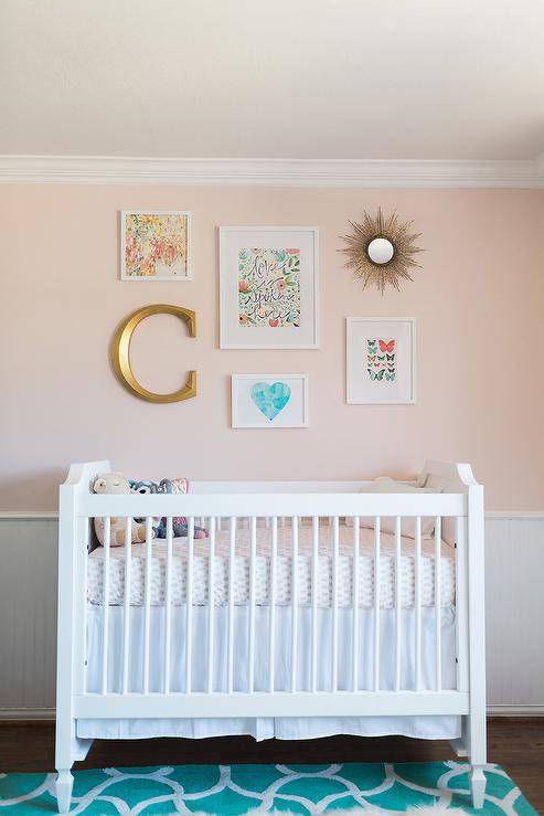 Placed on a green trellis rug, a white French crib is positioned beneath gallery art hung from a pale pink wall lined with white beadboard trim.