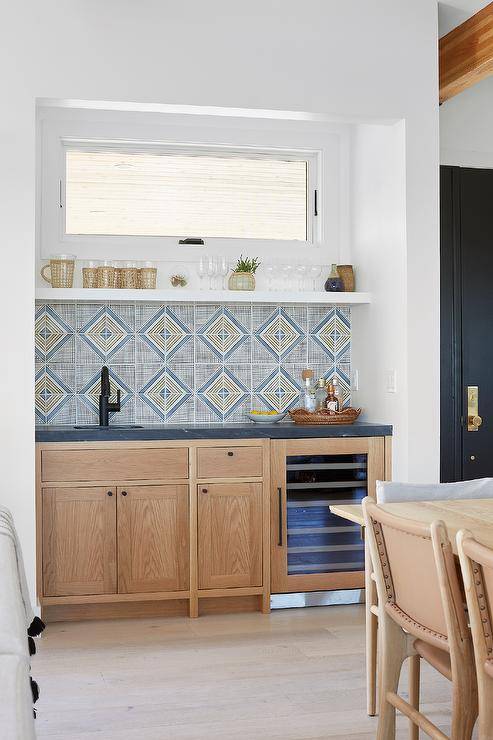 Small dining room wet bar boasts light brown oak cabinets donning small matte black knobs and a black marble countertop. The countertop is fitted with a small sink and matte black faucet fixed beneath a white shelf located under a window and in front of gold and blue geometric backsplash tiles.