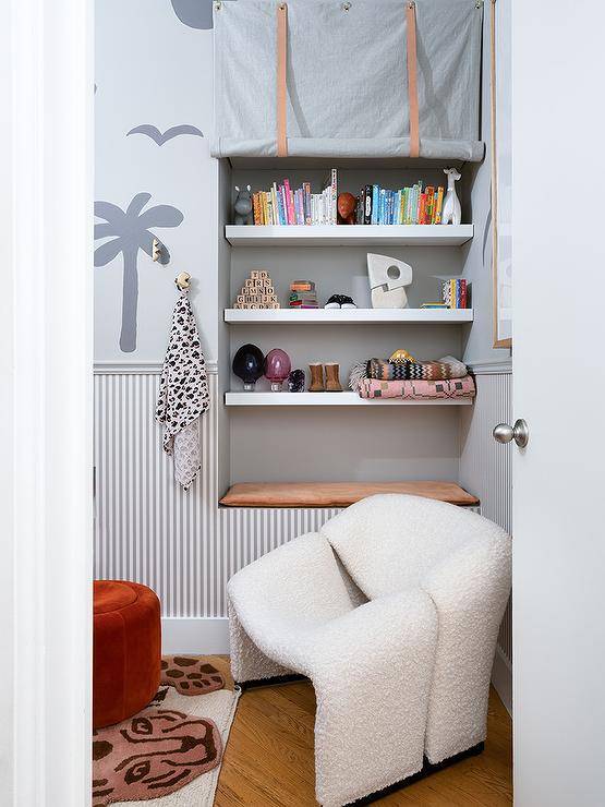 A white boucle chair sits in a modern nursery next to an recess fitted with a tan cocoon located underneath white shelves covered in a gray rolling curtain. A gray chair rail separates gray pinstripe wallpaper and gray gray patterned wallpaper.