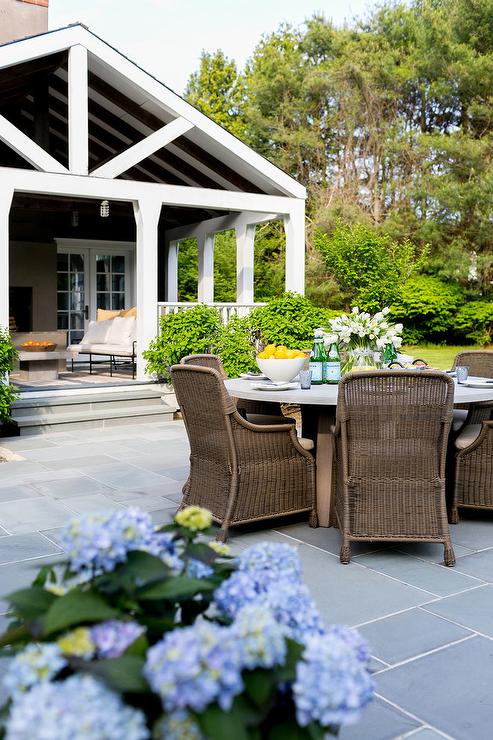 Slate patio paver floors showcasing a concrete dining table in front of a cottage. White frame cottage patio displays neutral furnishings under a vaulted ceiling.