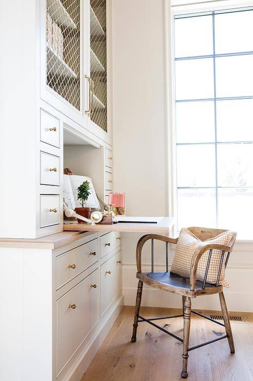 Add a rustic vintage wood chair a white built-in drop down desk for a well-balanced office space. Antique brass hardware accent the white inset cabinets and drawers along with brass metal grille doors. A large office window delivers ample light to showcase the sleek built-in finishings warmed by oak wood floors.