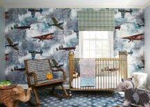 Nursery features Rebel Walls aero show wallpaper with vintage airplanes, a vintage black and white stripe nursery rocker, a brass crib, a gray rocking horse, a wooden elephant accent table atop blue and gray stars nursery carpeting and a green plaid window shade.
