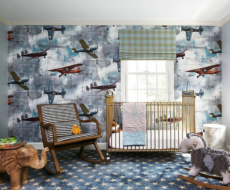 Nursery features Rebel Walls aero show wallpaper with vintage airplanes, a vintage black and white stripe nursery rocker, a brass crib, a gray rocking horse, a wooden elephant accent table atop blue and gray stars nursery carpeting and a green plaid window shade.