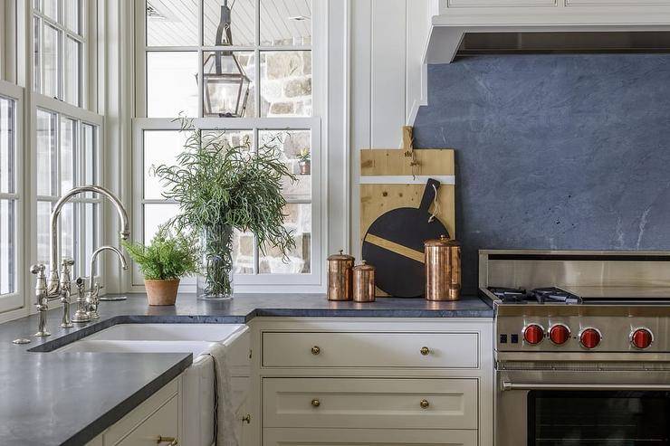 A Wolf range sits beside white kitchen cabinets contrasted with a black marble countertop and against a black marble slab backsplash complementing white shiplap wall trim. Windows are located above a dual farmhouse sink with a polished nickel gooseneck faucet.