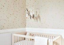 Blond wooden nursery crib in a nursery fitted for a girl or a boy features Buff & Gold Stars wallpaper, a white nursery crib an a feathered mobile.