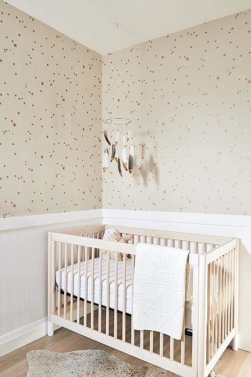 Blond wooden nursery crib in a nursery fitted for a girl or a boy features Buff & Gold Stars wallpaper, a white nursery crib an a feathered mobile.