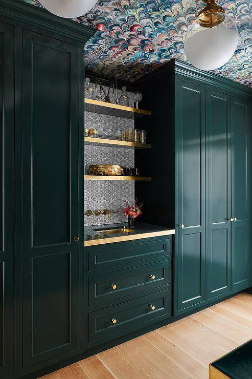 Fixed against gray mosaic marble backsplash tiles, brass shelves are fixed between glossy green lacquered floor-to-ceiling cabinets and over green shaker cabinets adorned with brass knobs and a brass countertop. The countertop is finished with a brass sink positioned beneath a wall-mount antique brass faucet.