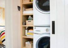 A hallway closet laundry room boasts a white sliding door with oil rubbed bronze hardware and a stacked white front loading washer and dryer fixed beside built-in oak shelves.